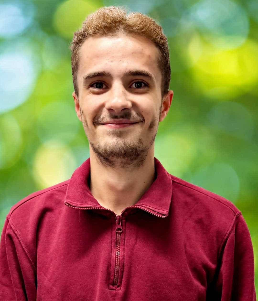 Porträt eines lächelnden jungen Mannes mit kurzem braunem Haar und rotem Sweatshirt vor grünem Hintergrund.
