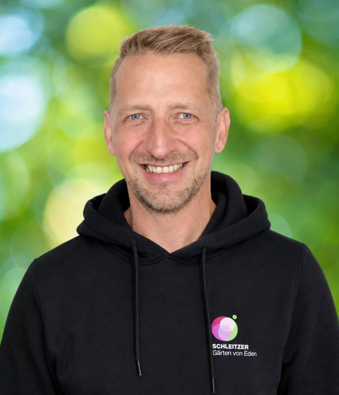 Juergen im schwarzen Schleitzer-Hoodie mit Gärten-vor-Eden-Logo Freundlicher Juergen mit kurzem blondem Haar und schwarzem Schleitzer-Hoodie vor grünem, unscharfem Hintergrund