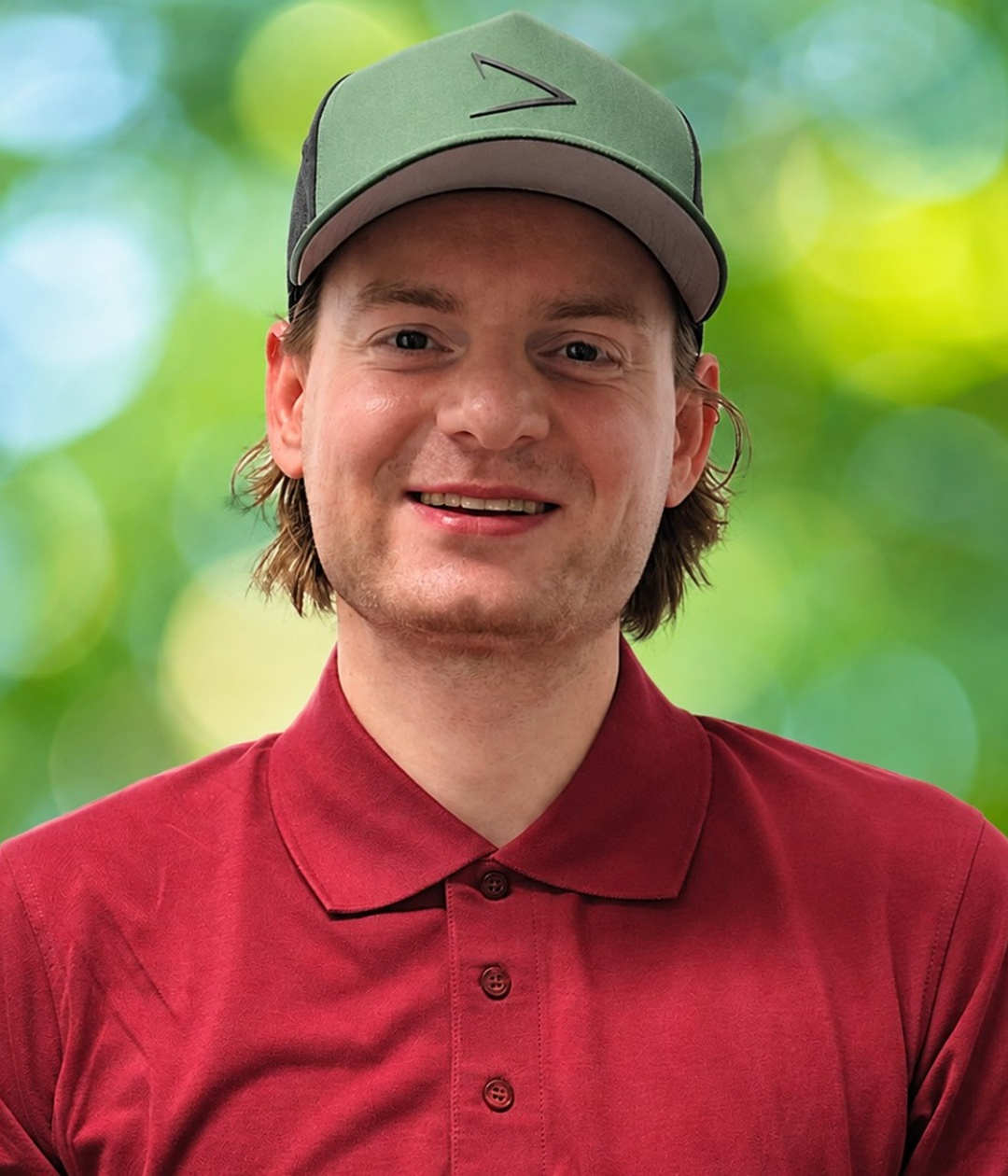 Linus im roten Poloshirt mit grüner Kappe im natürlichen Porträt Vor grünem Blattwerk lächelt Linus in die Kamera und trägt ein rotes Poloshirt mit grüner Kappe.