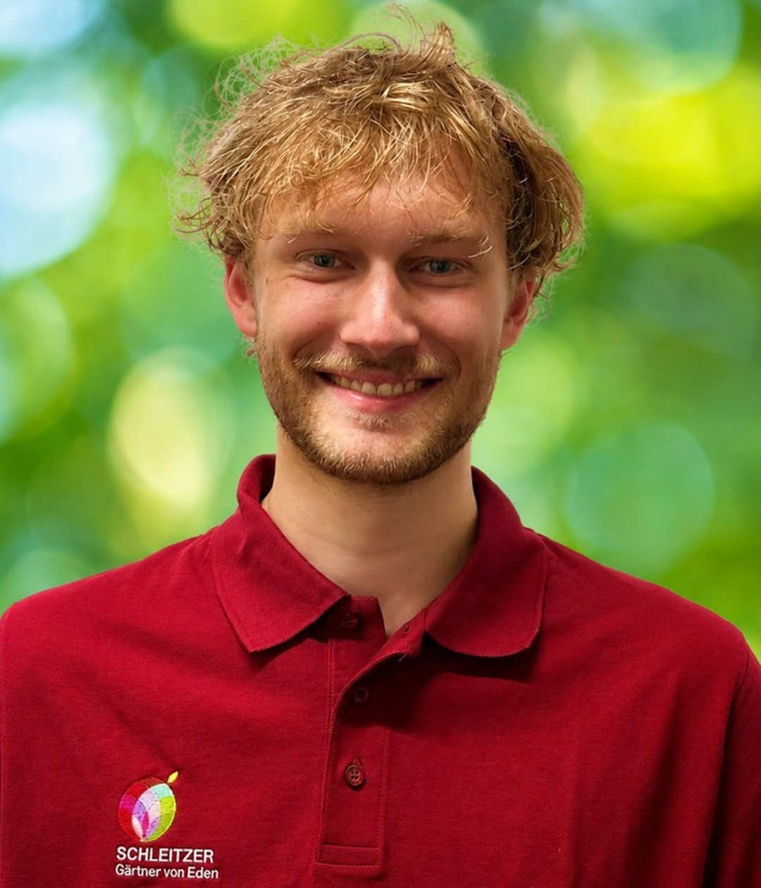 Mitarbeiterporträt von Schleitzer Gärtner von Eden im Grünen Ein lächelnder Mitarbeiter von Schleitzer Gärtner von Eden trägt ein rotes Poloshirt vor grünem, unscharfem Hintergrund.