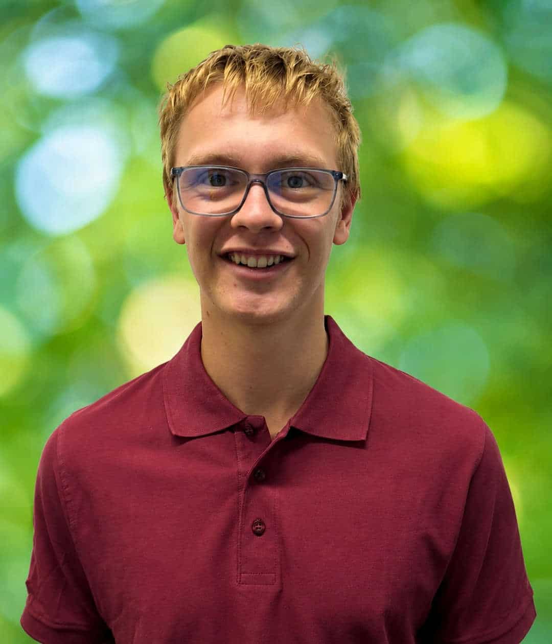 Simon_Rieger_1080x1260 Simon Rieger lächelt mit Brille und bordeauxrotem Poloshirt vor einem weich verschwommenen grünen Hintergrund.